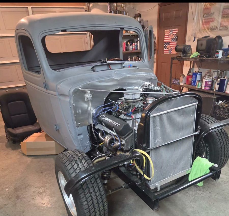 A 1930s Chevrolet pickup mid build in a garage, bare steel cab, small block Chevy engine with chrome covers, aluminum radiator and a four bar front end