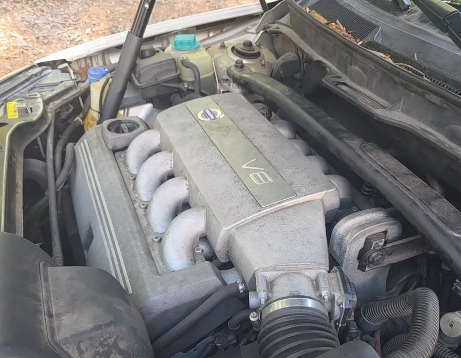 A Volvo V8 engine bay with the hood up, photographed outdoors on a leaf covered driveway